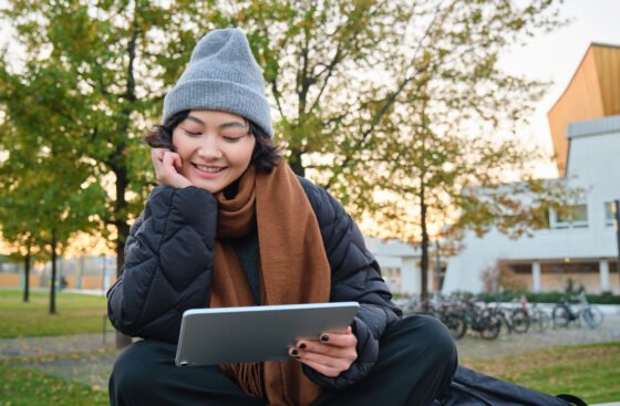 Portrait of asian girl relaxing in park, watching videos or reading on digital tablet, sitting on bench in hat and scarf on chilly day, enjoing outdoors.
